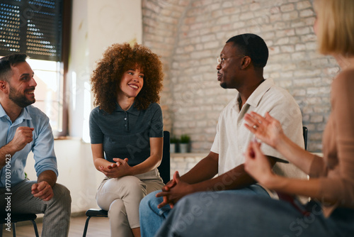 Smiling young woman with curly hair looks at an African American man speaking expressively during a positive and supportive multi-ethnic group therapy session indoors.