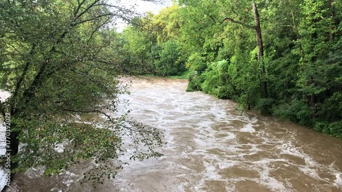 Rock Creek at flood stage in Washington, D.C., after heavy rains. The swollen, muddy brown river rushes quickly past the lush green trees and forested banks of Rock Creek Park.