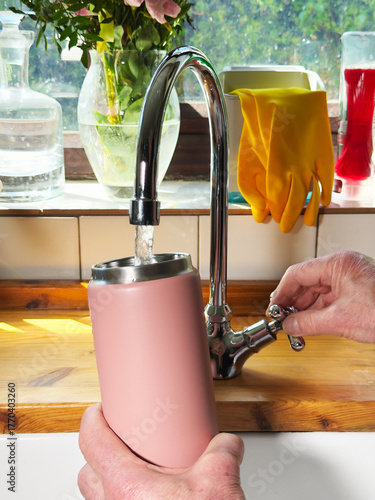 Closeup of one hand turning a kitchen sink tap, with the other holding an insulated drinks bottle while filling it with fresh running water.