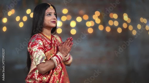 A beautiful Hindu woman wearing a red saree and gold ornaments stands gracefully with folded hands, offering prayers with a soft, blurred background.