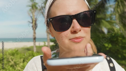 Smiling young woman wearing sunglasses and a bandana, holding a smartphone near her mouth, recording a voice message or talking on loudspeaker during a sunny beach vacation