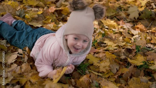 A medium-length shot of a little girl in a warm sweater and jeans sitting amidst a thick carpet of fallen leaves. The child is cheerfully holding a handful of colorful leaves in her hands.