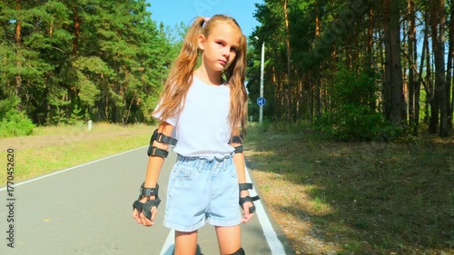 Child girl in casual clothes roller skating on road in forest on sunny summer day.
