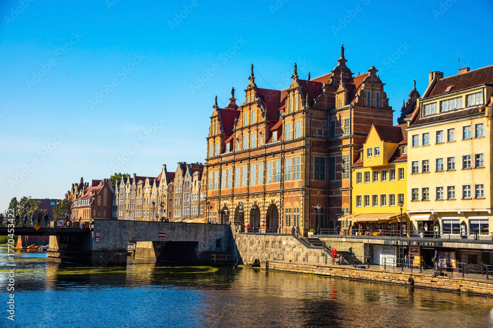 Fototapeta premium Scenic view of historic architecture and a bridge over a calm river in Gdansk, Poland. Bright day showcases ancient buildings reflecting in the water, offering cultural and travel allure.