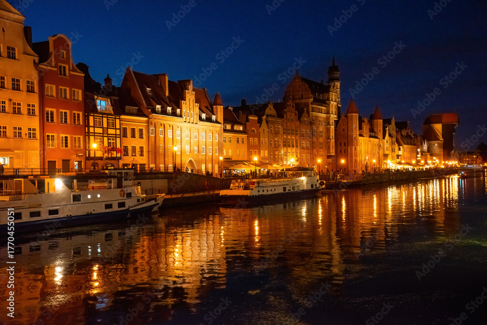 Obraz premium Gdansk riverside at night showcases historic buildings beautifully lit, reflecting on Motlawa River. People stroll along the promenade, enjoying a peaceful evening atmosphere