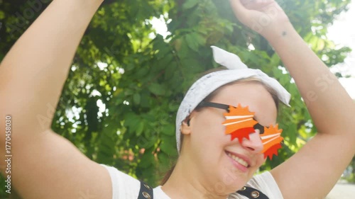 Young woman happily dancing and celebrating with a takeaway coffee cup in her hand, wearing quirky starburst sunglasses and a bandana, enjoying a vibrant summer day outdoors