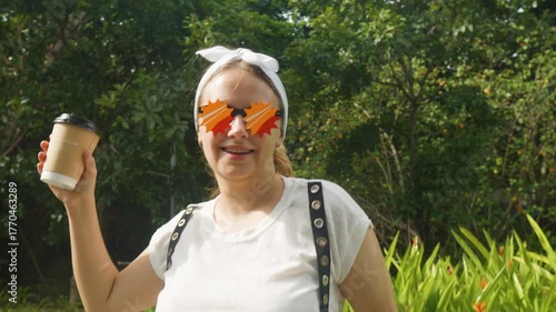 Young woman smiling while holding a takeaway coffee cup, wearing novelty star sunglasses and a white headband, standing in a lush green park enjoying a cheerful and casual moment outdoors