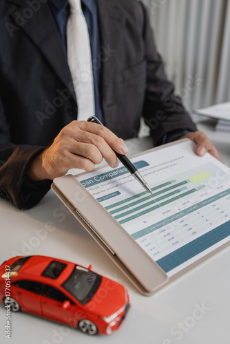 A businessman in a suit is calculating loan details, reviewing financial documents, and holding car keys near a calculator and toy car models, representing auto loan and finance services.
