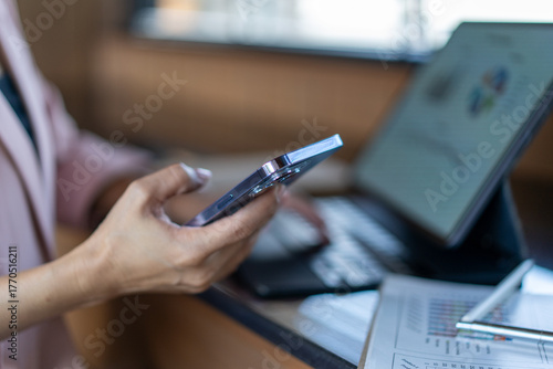 A woman is holding a cell phone and typing on a laptop