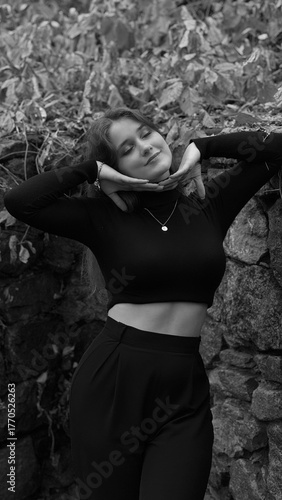 A woman in a black crop top and wide-leg pants stands by a stone wall on the steps in a park. She has her head thrown back and is relaxedly breathing in the fresh air, enjoying a moment of peace.