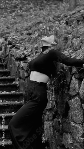A woman in a black crop top and wide-leg pants stands by a stone wall on the steps in a park. She has her head thrown back and is relaxedly breathing in the fresh air, enjoying a moment of peace.