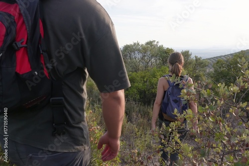 A couple with backpacks walks downhill along a natural trail surrounded by bushes and trees, illuminated by soft evening sunlight.