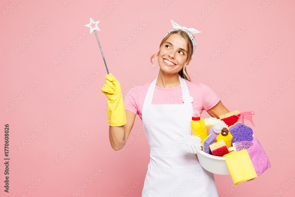 Obraz premium Young woman wearing white apron hold basin with detergent bottles while doing housework tidy up use magic wand fairy stick point aside on area isolated on plain pink background. Housekeeping concept.