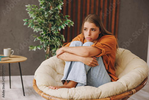 Full body young sad depressed frustrated woman wear orange casual clothes hold knees sits in chair stay at home hotel flat rest relax spend free spare time in living room indoor. People lounge concept