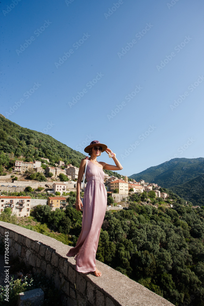Naklejka premium Beautiful elegant woman in a light rose dress walking gracefully in front of the spectacular view of Cargèse village Corsica, France. Scenic Mediterranean landscape with charming coastal architecture