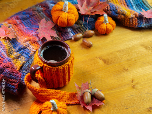 Colorful autumn composition, a mug of hot coffee in a knitted cover on a wooden table. Knitted small pumpkins and a scarf. Copy space