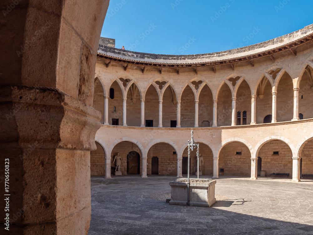 Obraz premium Castell de Bellver. Palma de Mallorca, historic circular courtyard features a stone fountain in the center, surrounded by arched colonnades with a clear blue sky overhead, Majorca