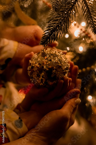 children's hands holding a Christmas tree ornament in front of a Christmas tree at night