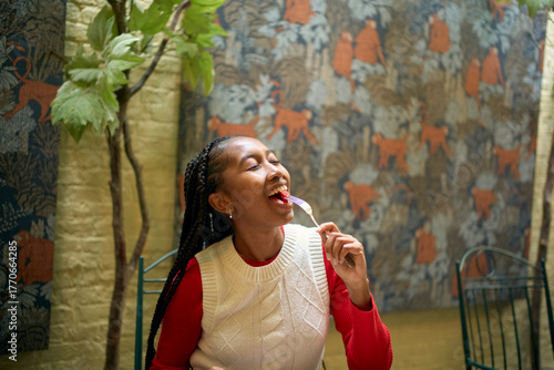 Woman in a red top and white vest eats a treat in a patterned room with a tree. Bruges, Belgium