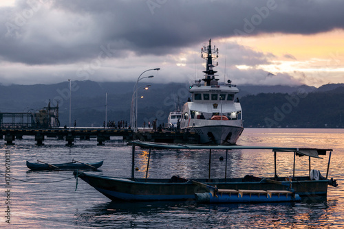 A large docked ship at dusk with a smaller boat in the foreground and cloudy mountains beyond. Ambon, Maluku, Indonesia