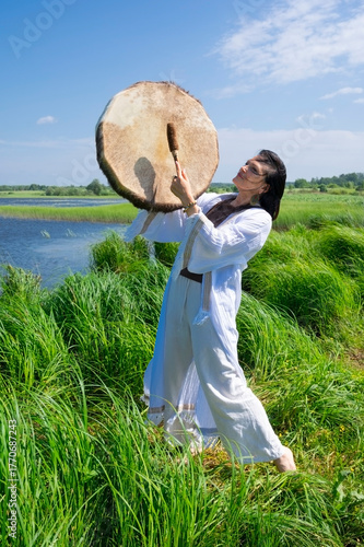 Female shaman in a trance in the white dress drumming in the natural environment