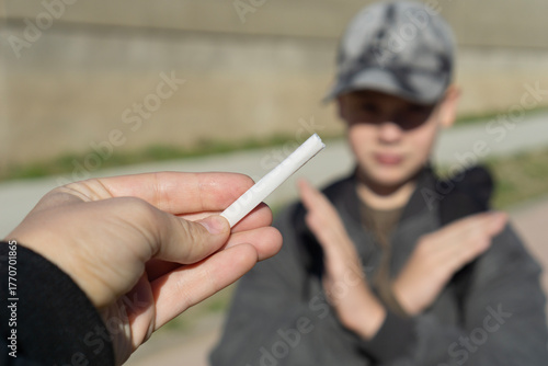hand offering a white cigarette to a young boy in a cap who is blurred and making a clear no, stop gesture with his arms, concept of youth health, antismoking campaign, education