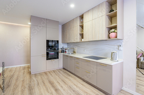 A modern kitchen with light wood flooring, neutral-toned cabinets with gold handles, built-in appliances, and open shelving. The design is clean and minimalist