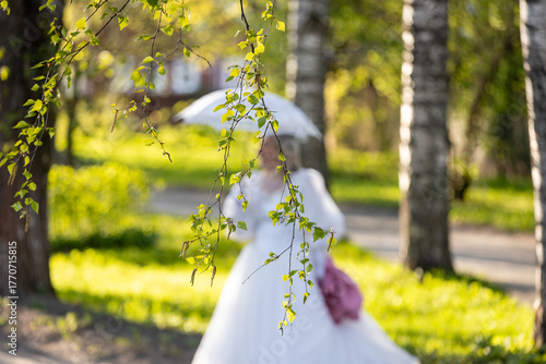 Romantic bride in white wedding dress holding umbrella, walking through sunlit spring park with birch trees, soft bokeh background — perfect for love, marriage, and nature-themed concepts.