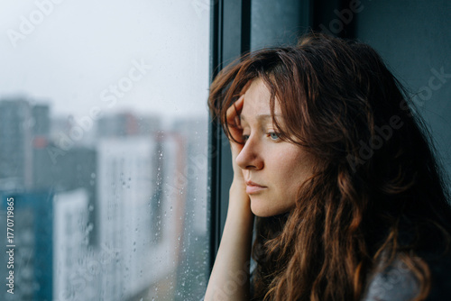 Young woman feeling sad and pensive, experiencing autumn depression while looking out a rainy city window, pondering loneliness and mental health challenges during a melancholic season