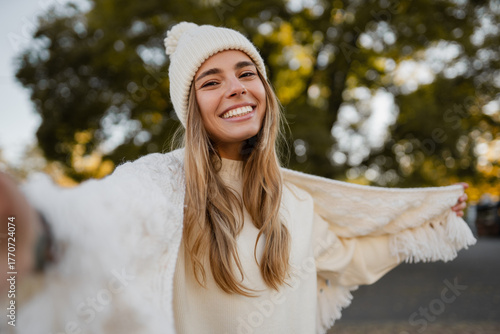 attractive smiling young blond woman walking in winter park having fun in warm white knitted sweater, cape and hat, cold season