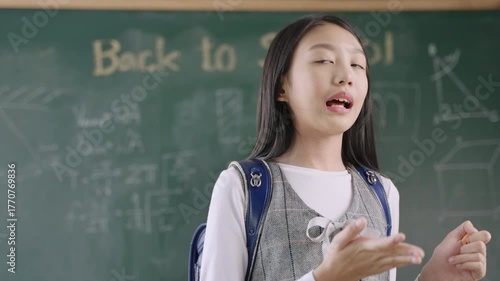 Back to school concept. Portrait of happy Asian child girl student in uniform with a school bag and backpack standing in front of a blackboard with a myself speech and presentation in the classroom. 