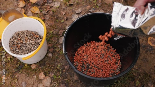 Preparing fishing groundbait with pellets and additives in bucket, close-up of angler mixing ingredients for carp fishing on autumn ground, detailed outdoor hobby and bait preparation concept