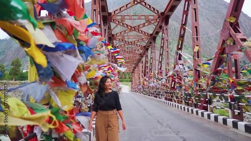  Indian girl walk near colorful prayer flags hanging on metal bridge in mountain valley near Mini Manali in Lahaul, Himachal Pradesh, India. Female tourist exploring remote village of Himalayas. 