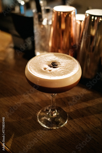 Espresso Martini (Cocktail) mit 3 Kaffeebohnen (bringen Glück) auf Holzbar, dahinter Cocktailshaker (unscharf)