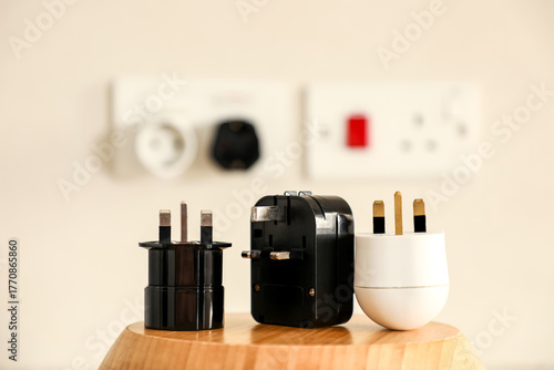 On a wooden surface lie one white and two black travel adapters, with blurred international sockets in the background. Minimalist composition highlighting global connectivity and modern travel.