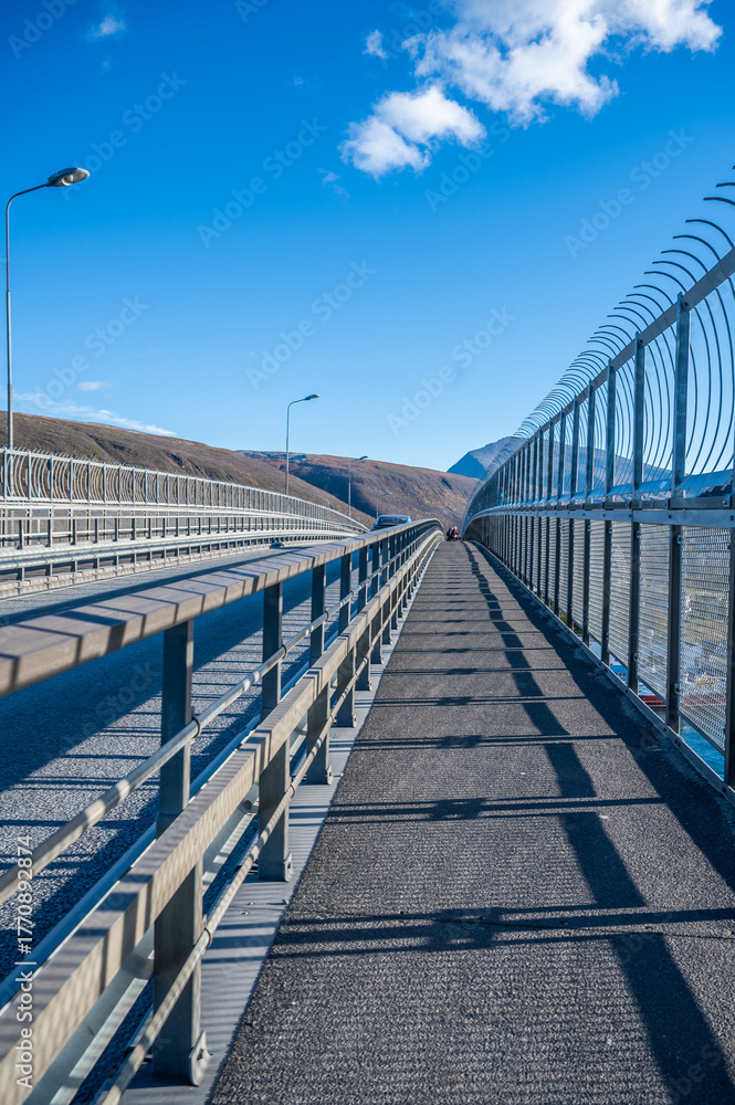 Naklejka premium Tromsobrua Bridge long, empty bridge with a metal railing and barbed wire on the right, set against a clear blue sky and distant mountains, Tromso Norway