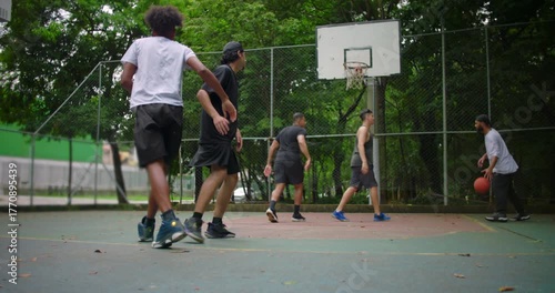 Hispanic man helping friend of African descent up from ground after fall on outdoor basketball court, teammates reuniting to continue friendly street game