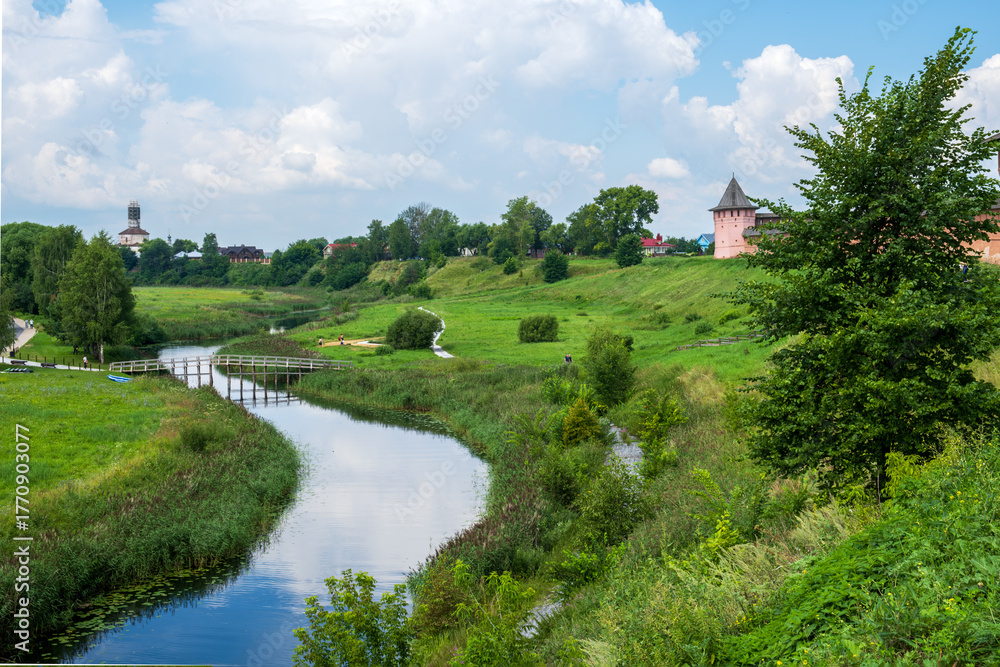 Obraz premium e landscape of the outskirts of a town in the Russian provinces, with a winding river, lush greenery, and a fortress wall in the distance, under a bright summer sky.