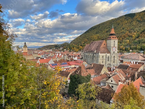 A panoramic view of a charming old town with red-tiled roofs, a historic clock tower, and a large gothic church surrounded by autumn trees and forested hills under warm afternoon sunlight.
