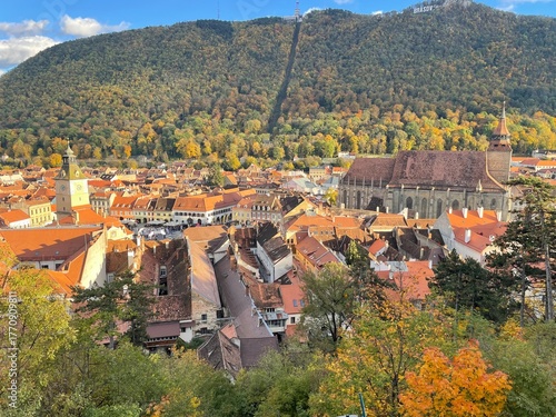 A panoramic view of a charming old town with red-tiled roofs, a historic clock tower, and a large gothic church surrounded by autumn trees and forested hills under warm afternoon sunlight.
