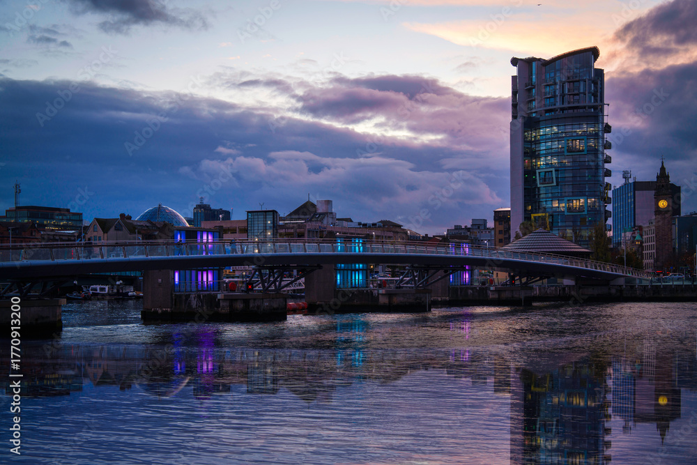 Naklejka premium Belfast City Skyline at sunset with dramatic clouds over the Lagan Weir Pedestrian and Cycle Bridge along the Lagan River in Northern Ireland, United Kingdom.