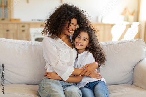 Loving Latin family mother and daughter bonding at home, woman and girl embracing, sitting on couch and smiling, copy space