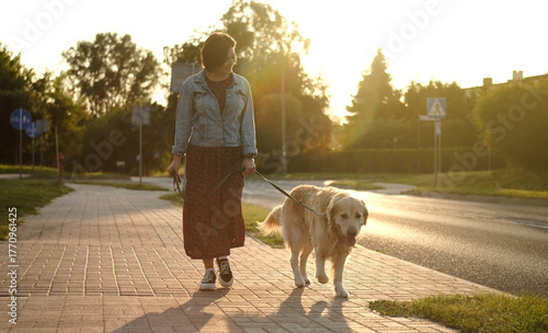Female Dog Owner Walks Adorable Golden Retriever At Sunset On A Street During Daily Routine