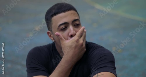 Close-up of Latin Hispanic man of African descent sitting outdoors, looking sideways with expression of inner conflict and restrained angst, lost in thought