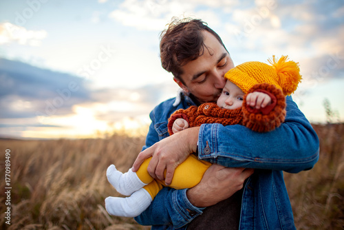Happy man kissing his little baby girl walking outdoors. Father holding infant daughter wearing fall brown sweater and yellow hat