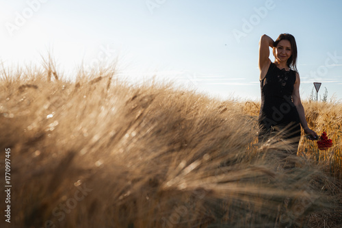 Elegant woman in a black dress in a golden wheat field at sunset, holding red flowers. Evokes rustic beauty, tranquility, and natural grace. Summer romance