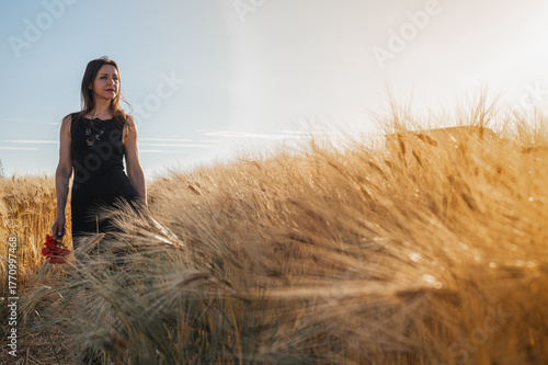 Elegant woman in a black dress in a golden wheat field at sunset, holding red flowers. Evokes rustic beauty, tranquility, and natural grace. Summer romance
