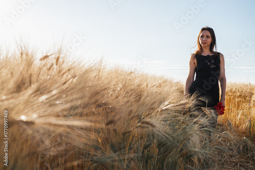 Elegant woman in a black dress in a golden wheat field at sunset, holding red flowers. Evokes rustic beauty, tranquility, and natural grace. Summer romance