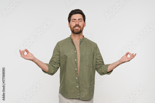 Young tranquil man in casual clothes standing isolated on white background makes mudra gesture meditates feels internal balance having healthy habit, wellbeing harmony no stress concept