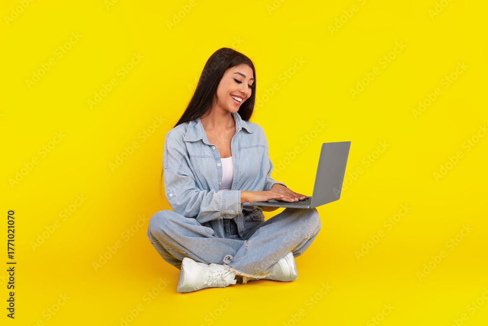 Naklejka premium A woman sits comfortably on the floor, legs crossed, with a joyful expression while typing on her laptop. The bright yellow backdrop adds a cheerful vibe to the scene.